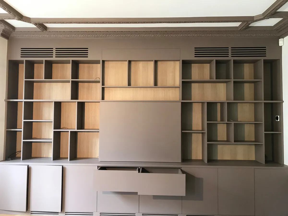 Après travaux : bibliothèque sur mesure en chêne et laque taupe occupant tout le mur, grilles de ventilation intégrées