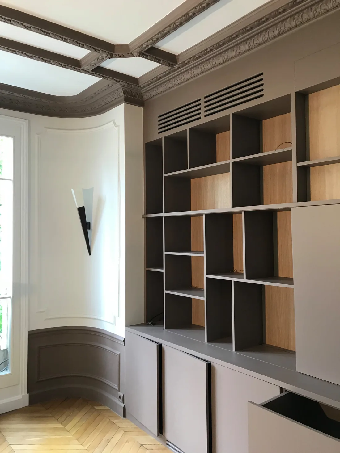 La bibliothèque vue depuis l'angle de la pièce : retour du meuble le long du mur en pan coupé, grilles de ventilation en partie haute et corniche ancienne conservée au plafond