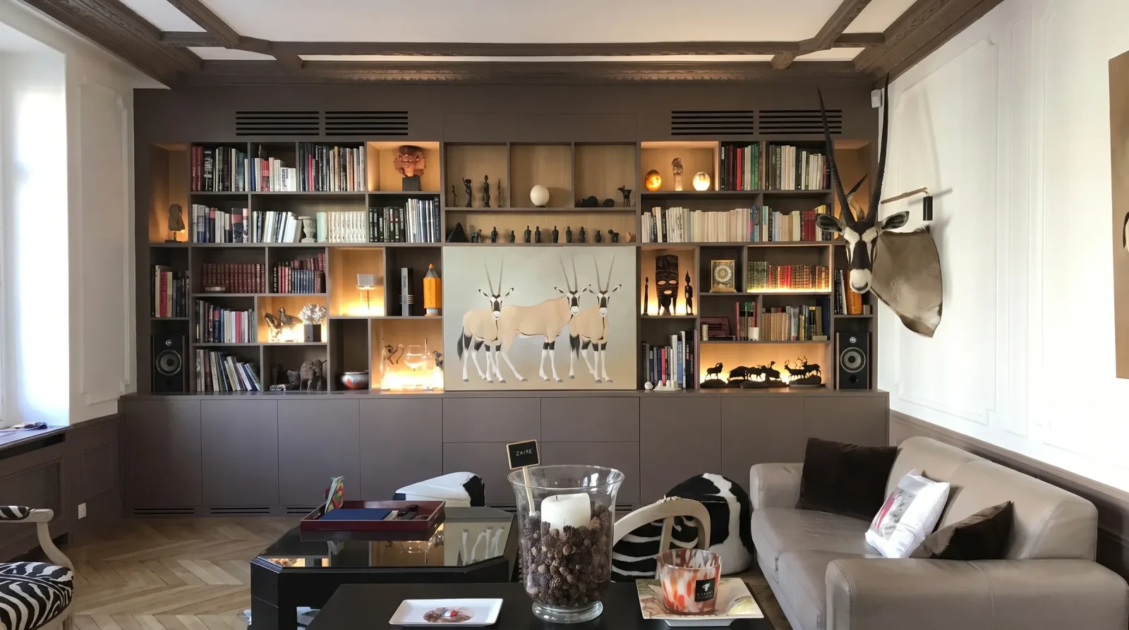 Le salon avec la bibliothèque sur mesure terminée : cases éclairées garnies de livres et d'objets, grande toile aux trois oryx posée devant le panneau central, enceintes intégrées de part et d'autre, plafond à caissons brun et parquet point de Hongrie