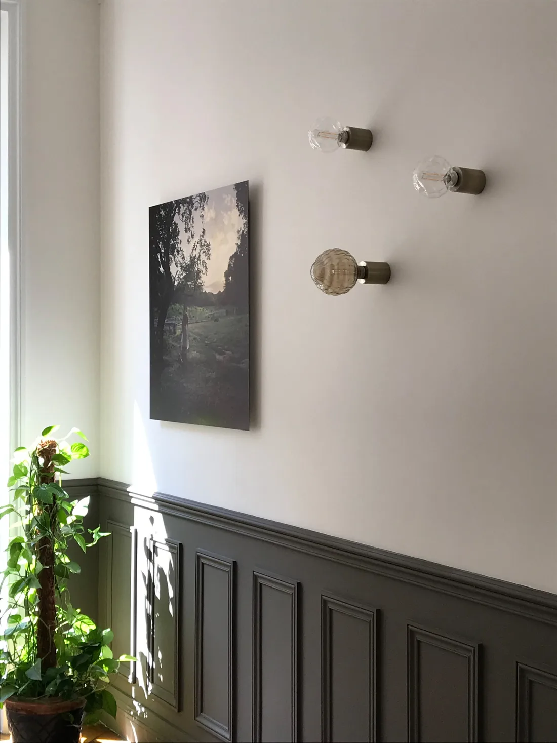 Trio d'appliques de laiton à globes de verre facetté et strié sur un mur blanc, au-dessus d'un soubassement de boiseries gris anthracite, à côté d'une photographie de paysage encadrée