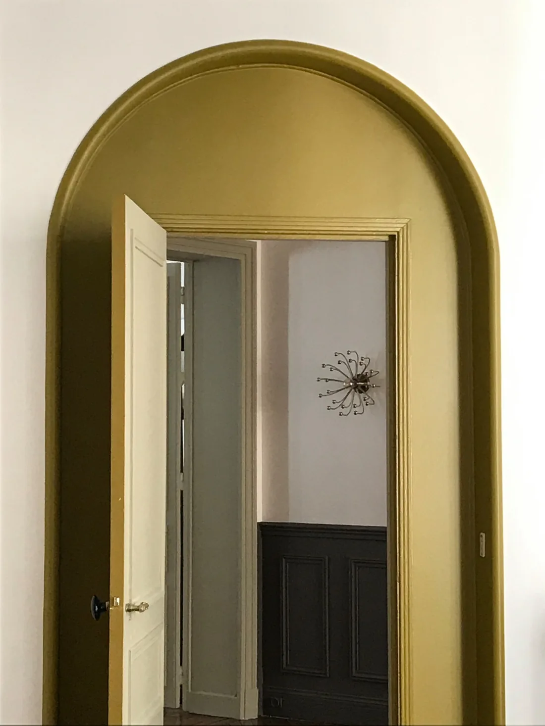 Arche de passage peinte en jaune moutarde dans toute son épaisseur, ouvrant sur une pièce au soubassement gris anthracite où est accrochée une applique étoilée dorée