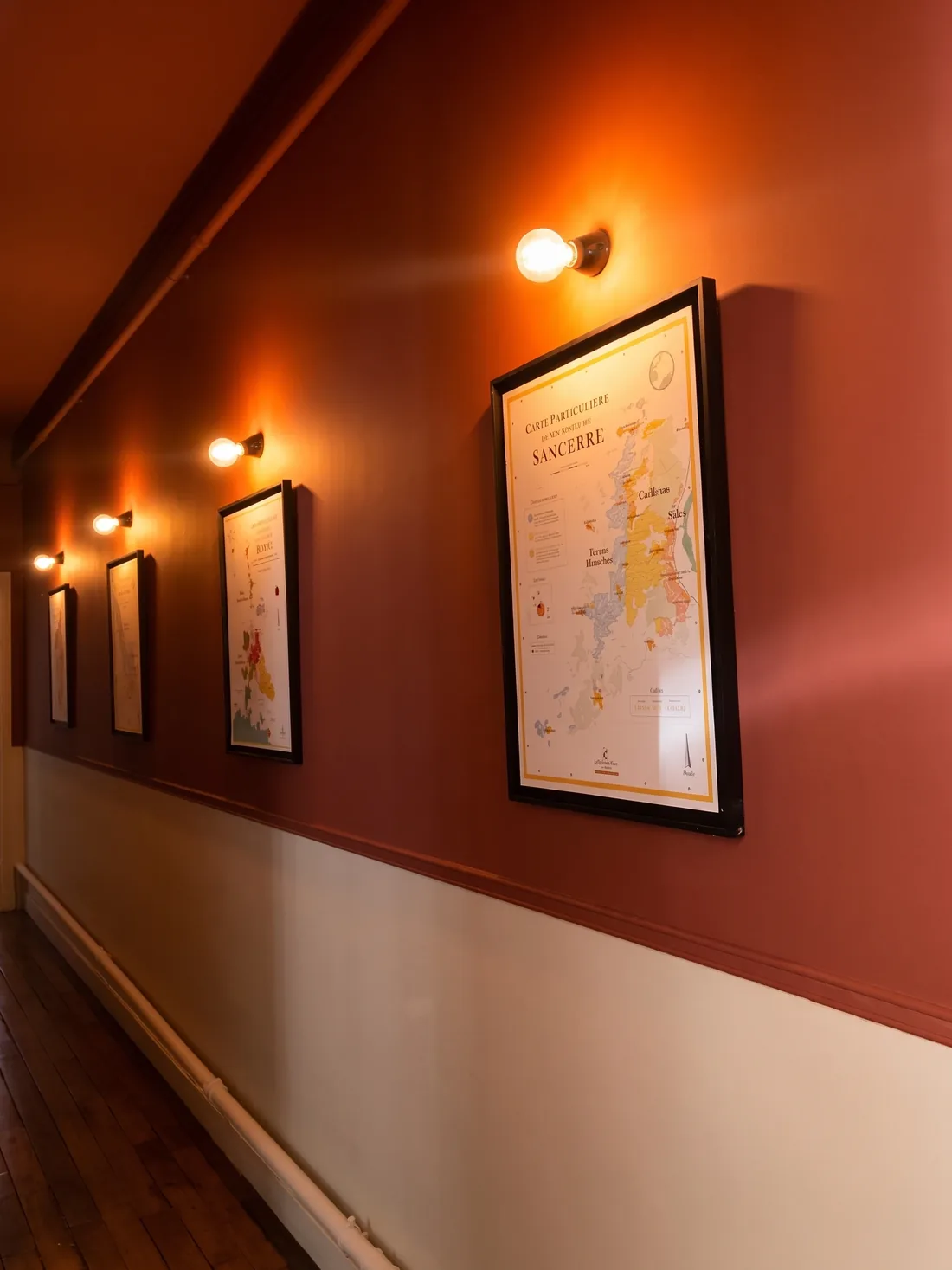 Le couloir après : murs terracotta profond, cartes de vignobles encadrées de noir accrochées en enfilade, chacune éclairée par une applique à ampoule nue, soubassement clair conservé