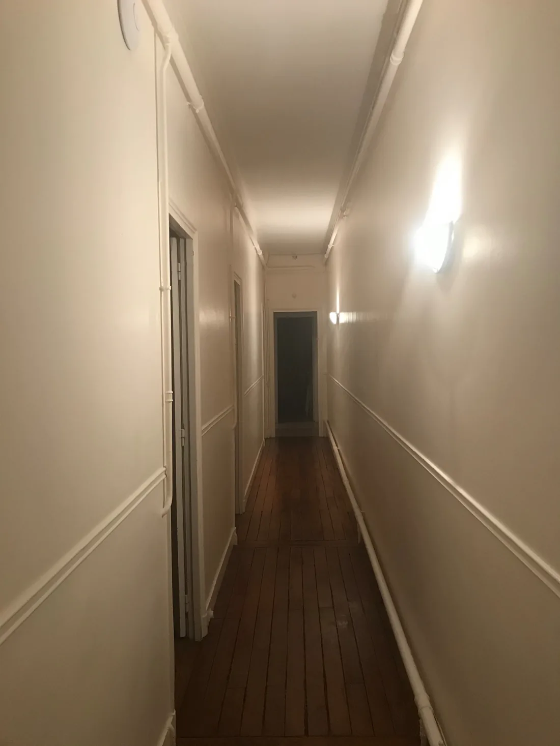 Le couloir avant : murs clairs sans décor, tuyaux apparents le long du plafond et de la plinthe, enfilade de portes et parquet ancien filant vers une porte sombre au fond