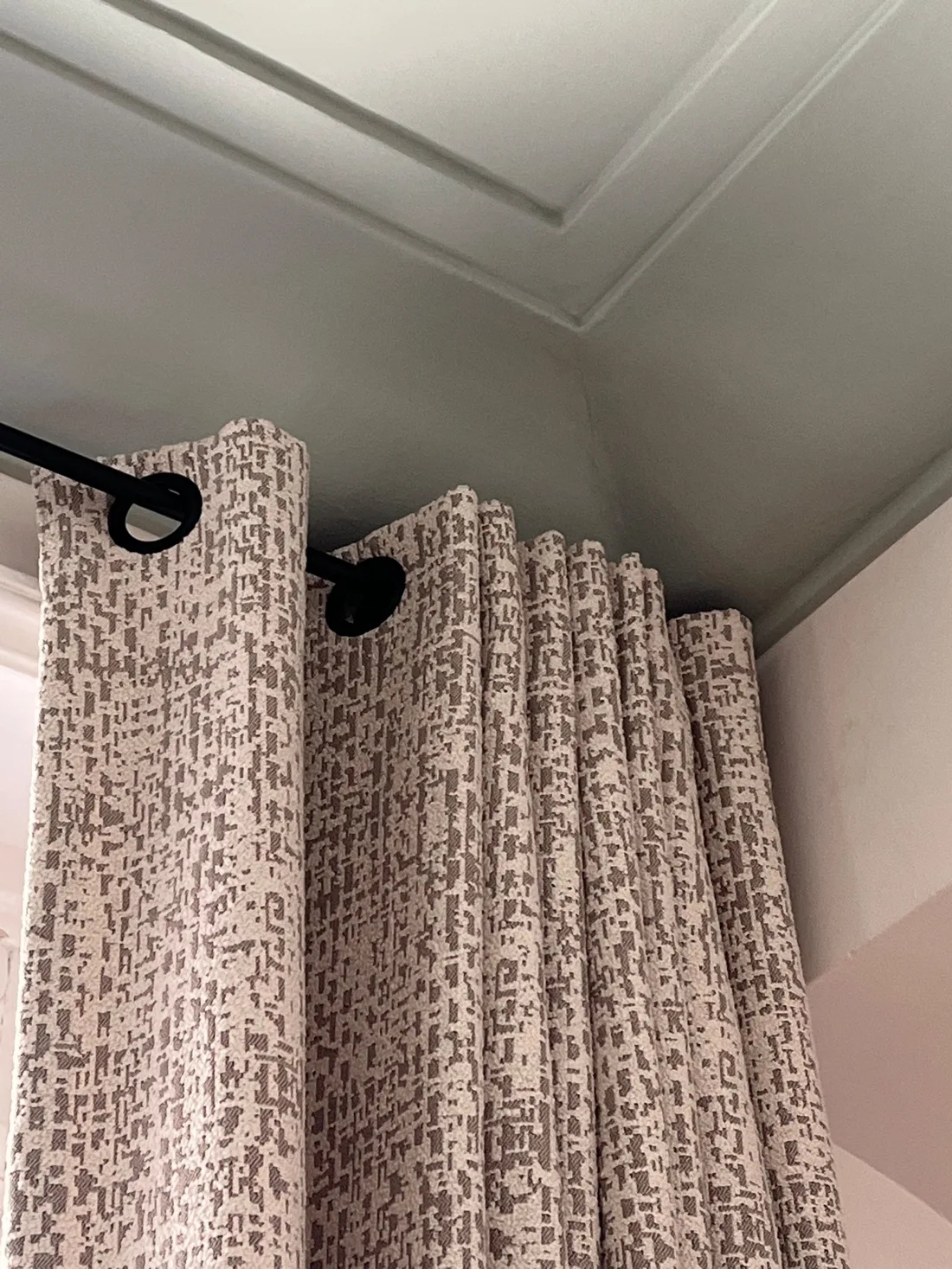 Détail des rideaux au tissage chiné beige et taupe sur leur tringle noire à œillets, sous la corniche du plafond peint en vert céladon