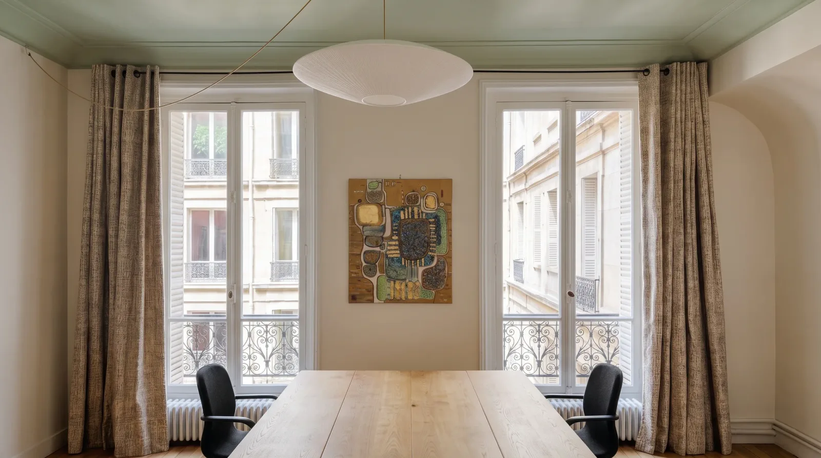 Salle de réunion dans un appartement haussmannien : plafond peint en vert céladon, grande suspension plissée blanche, table de bois clair, deux fenêtres à garde-corps ouvragés donnant sur la rue et tableau abstrait aux tons or et turquoise