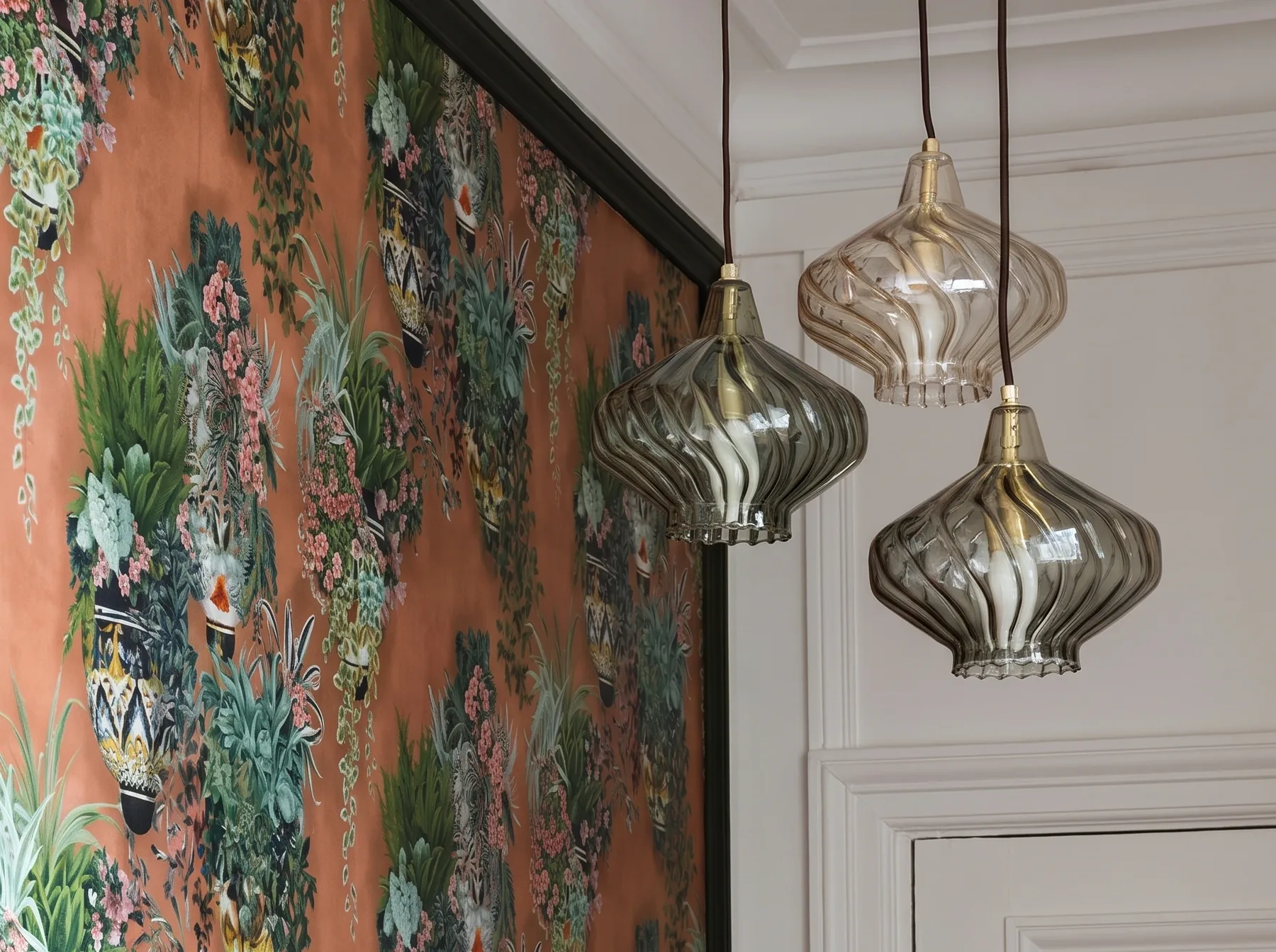 Panneau de papier peint à vases fleuris sur fond terracotta dans un encadrement vert sombre, au milieu de boiseries blanches, avec au premier plan trois suspensions de verre soufflé fumé et ambré à godrons torsadés