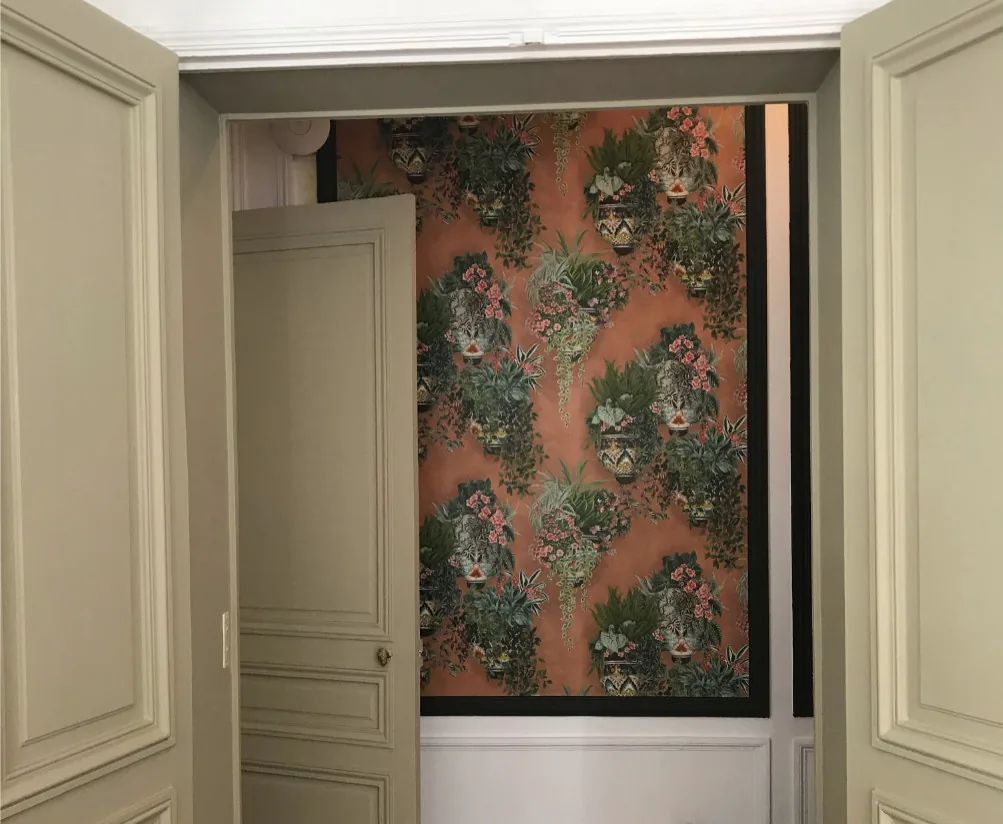 Porte à moulures peinte en gris vert ouverte sur le panneau de papier peint à vases fleuris, vu depuis la pièce voisine aux boiseries claires