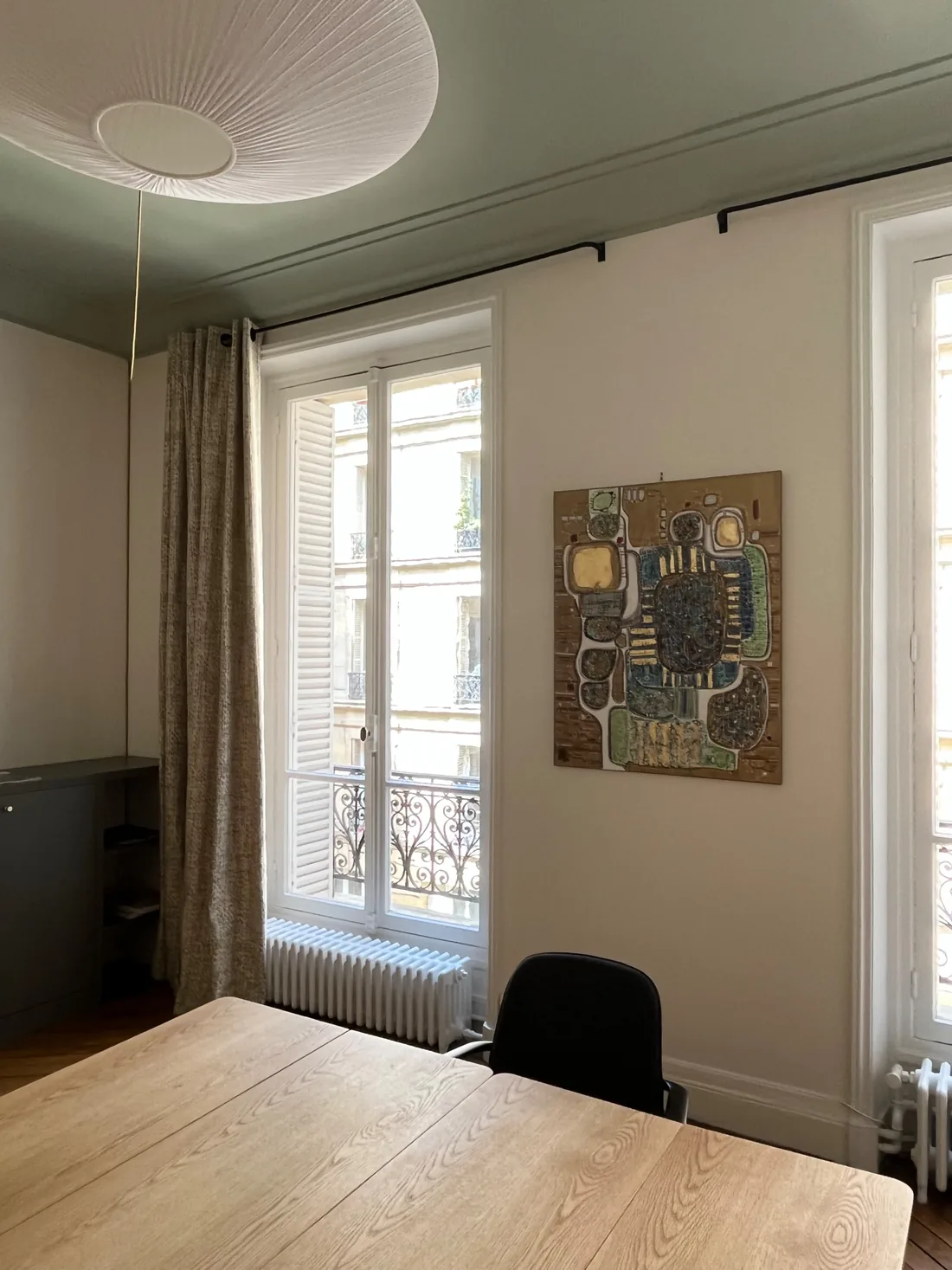Poste de travail près de la fenêtre : table de bois clair, chaise de bureau noire, radiateur en fonte sous la fenêtre à garde-corps et tableau abstrait aux tons or et turquoise au mur