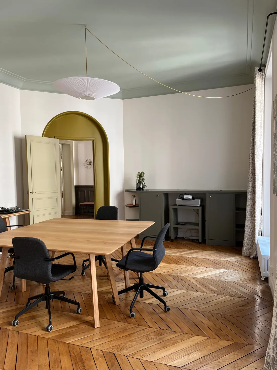 Salle de réunion vue en enfilade : parquet point de Hongrie, table de bois clair et fauteuils de bureau noirs, plafond vert céladon et arche jaune moutarde ouvrant sur la pièce voisine