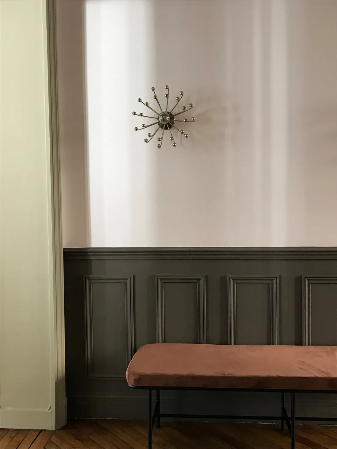Angle d'une pièce de travail : soubassement de boiseries peint en gris anthracite, banquette de velours rose à piètement de métal noir et applique étoilée dorée en hauteur sur le mur blanc