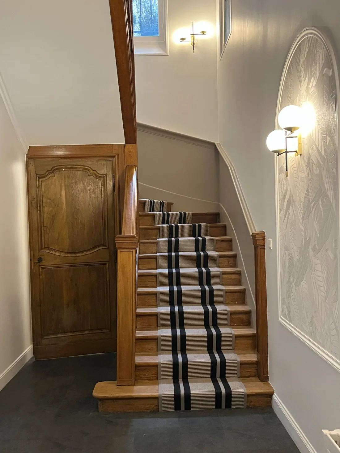 L'escalier : tapis à rayures graphiques noires et blanches sur les marches de bois clair, porte ancienne en chêne à droite, appliques à globes et panneau de papier peint texturé