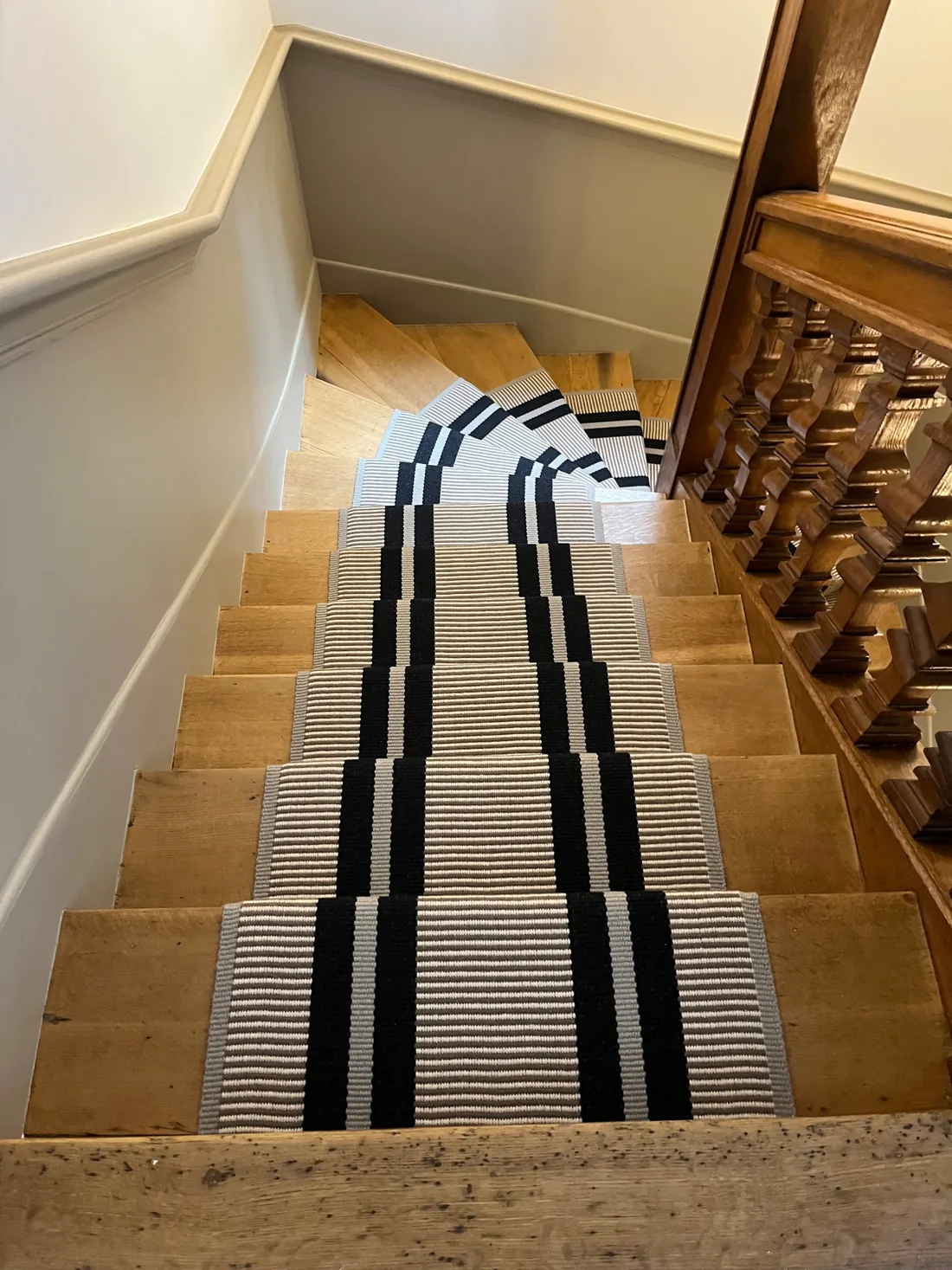 L'escalier vu en plongée : la rampe de bois tourné d'origine et le tapis à rayures noires et blanches épousant les marches