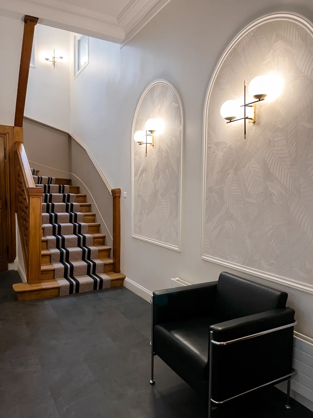 Le hall : panneaux muraux en arches revêtus de papier peint texturé, appliques doubles à globes lumineux, soubassement vert de gris, sol de marbre sombre et tapis d'entrée