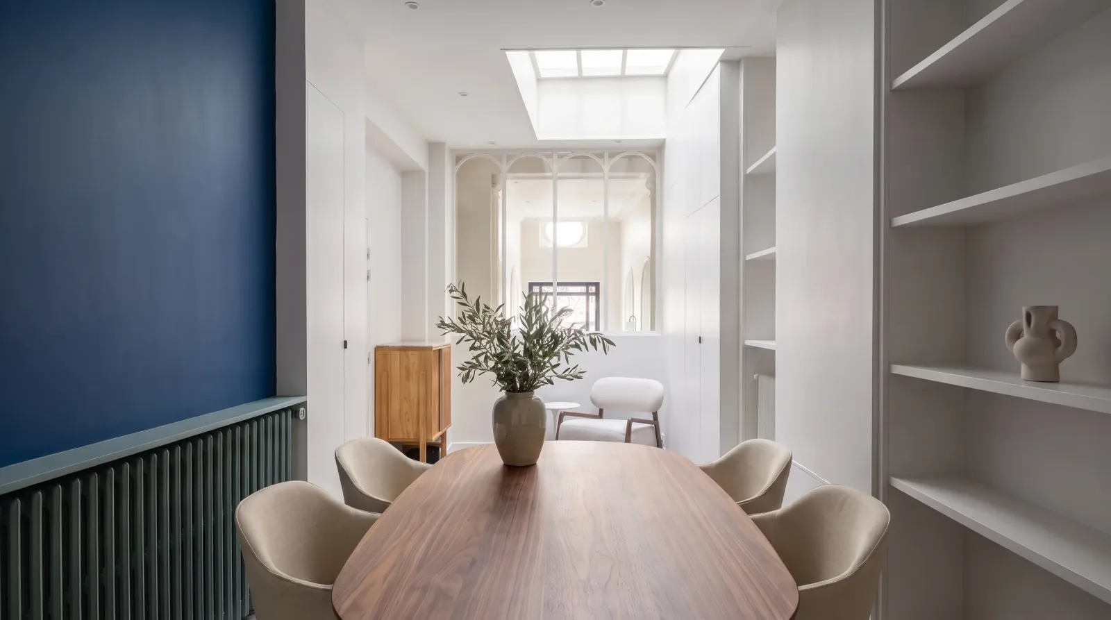 La salle de réunion des bureaux : table ovale en noyer entourée de fauteuils écrus, mur bleu nuit au-dessus d'un radiateur habillé vert de gris, puits de lumière au plafond, bibliothèque murale et verrière blanche en arches ouvrant sur le hall