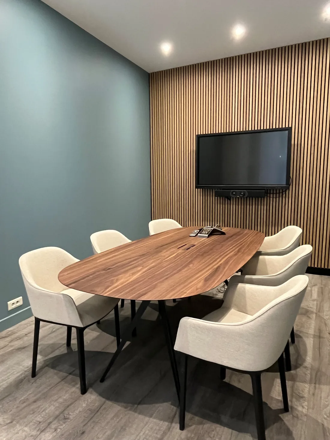La salle de réunion : table ovale en noyer, fauteuils écrus à piètement noir, mur de tasseaux de bois portant l'écran, murs vert de gris et spots encastrés au plafond