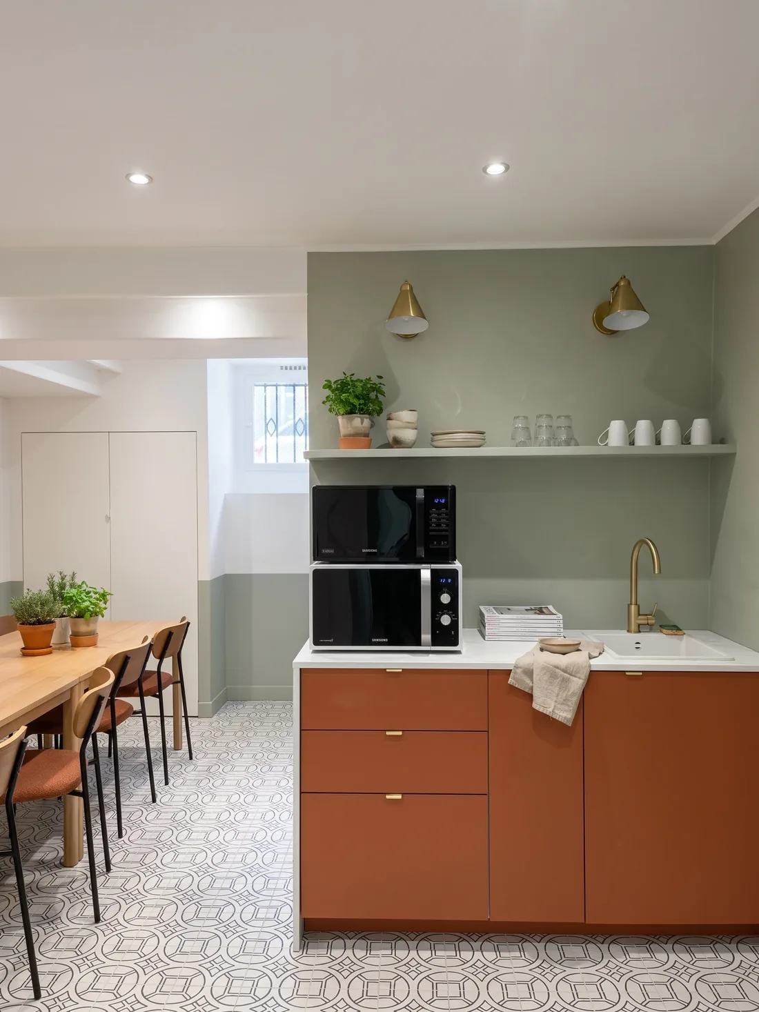 L'espace repas du sous-sol : meubles terracotta sous un plan blanc, crédence claire, appliques dorées, micro-ondes encastré et table de bois clair au premier plan