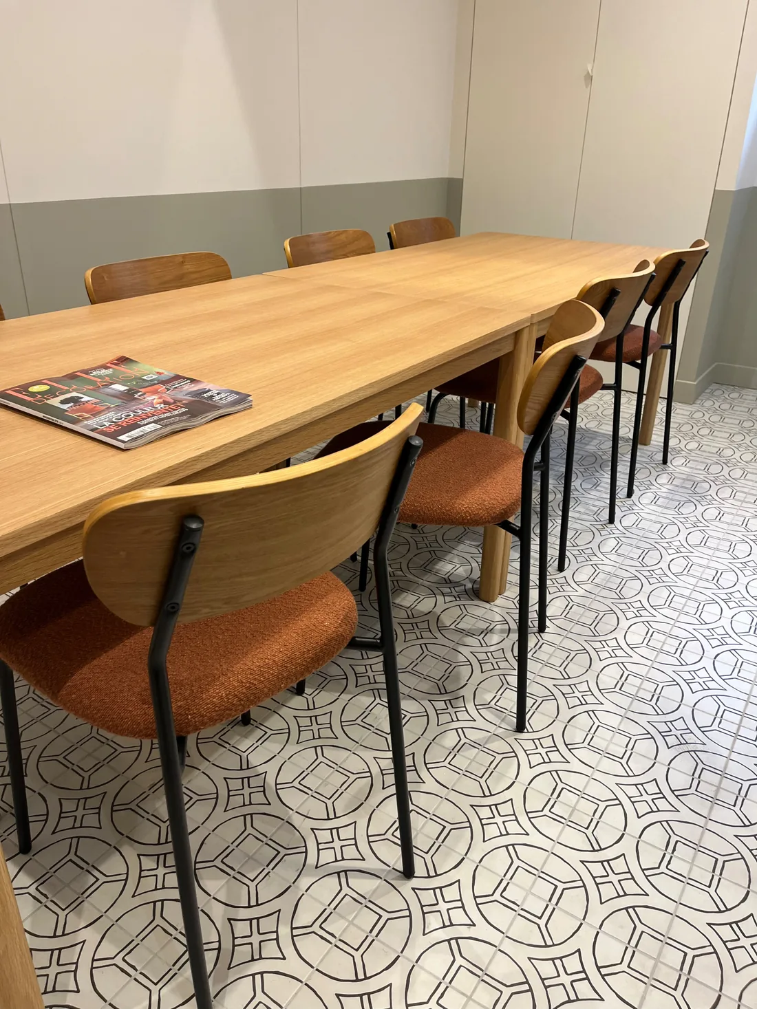 La grande table de réunion en chêne clair entourée de chaises aux assises cognac, sur un sol de carreaux graphiques gris et blancs
