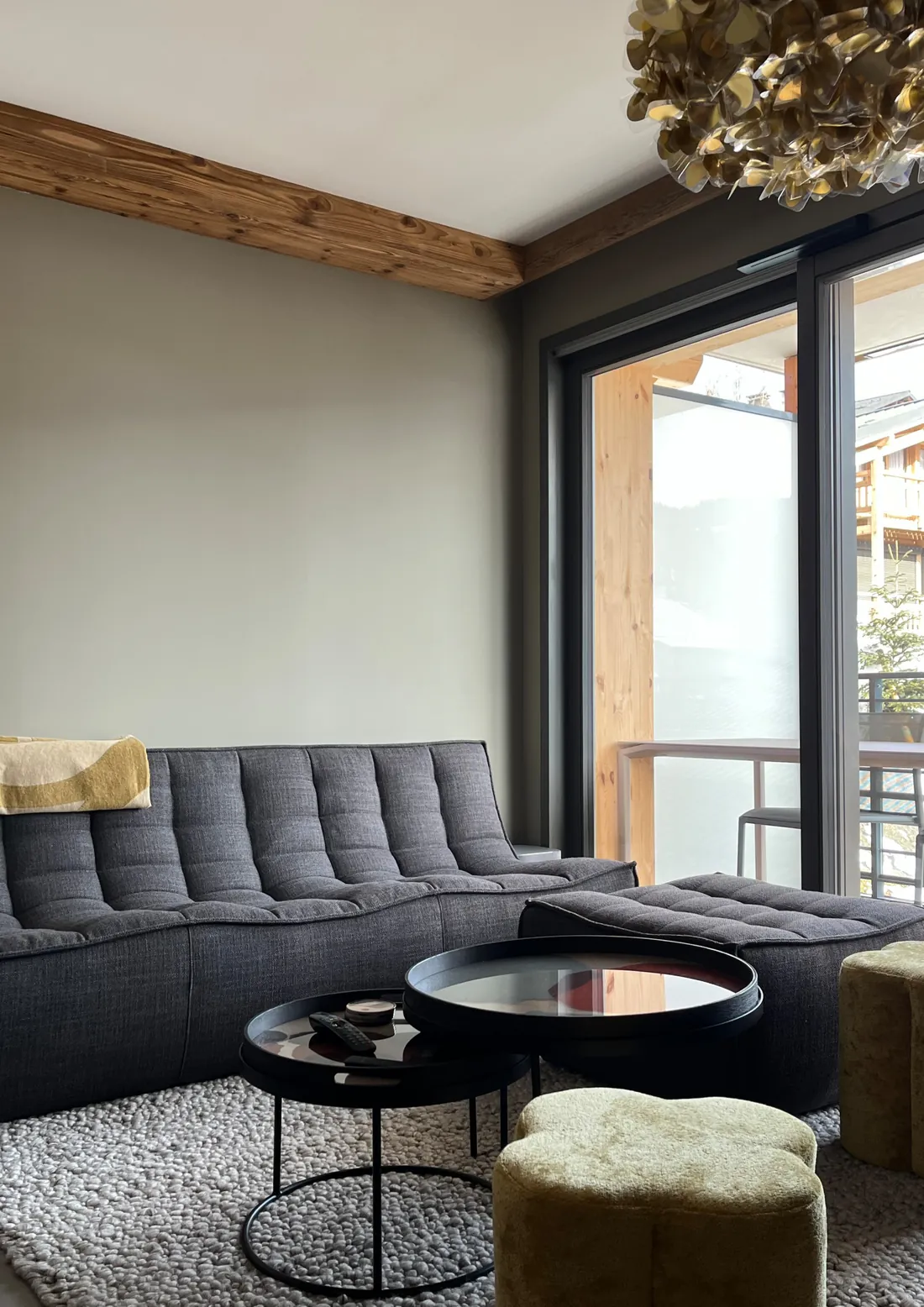 Canapé modulable gris à assises matelassées et repose-pieds, devant une grande baie vitrée coulissante donnant sur le balcon en bois, poutre apparente au plafond
