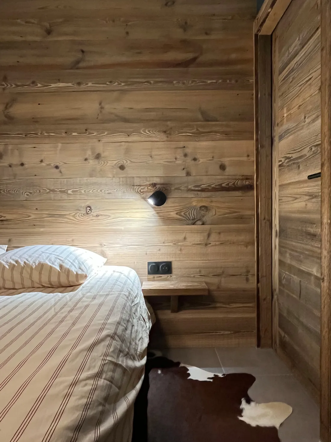 Détail de chambre : mur de lambris ancien, tablette de chevet suspendue en bois, liseuse orientable noire, linge de lit rayé écru et peau de vache au sol