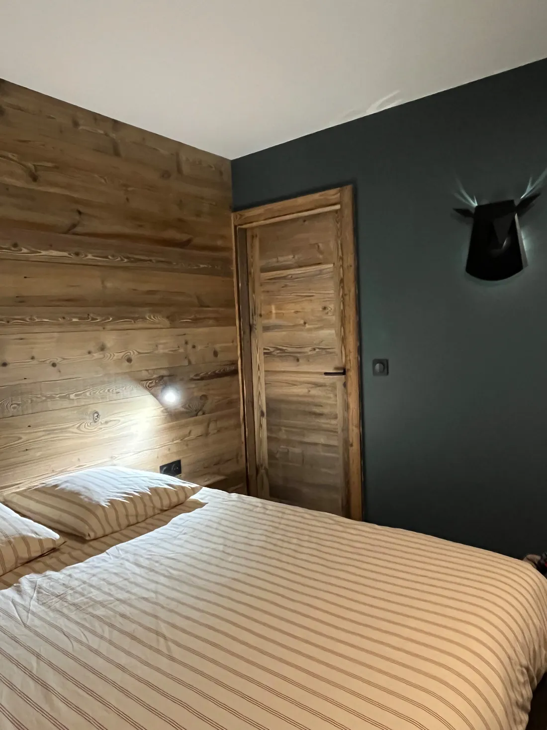 Seconde chambre : mur peint en vert sapin foncé, mur de tête de lit en lambris de bois clair, porte en vieux bois et applique murale noire en forme de tête de cerf stylisée