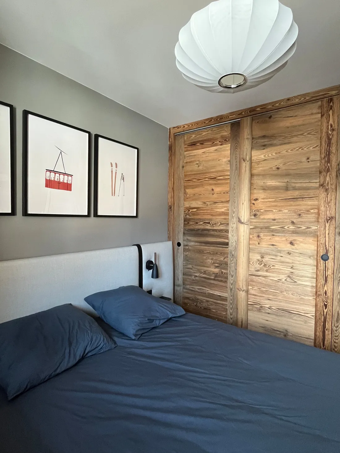 Chambre principale : lit en linge bleu nuit, tête de lit tendue de laine écrue, trois affiches de ski encadrées et grands placards à portes coulissantes en vieux bois