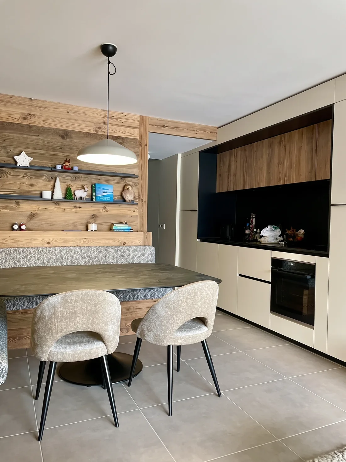 Vue d'ensemble de la cuisine et du coin repas : banquette d'angle sur mesure, tablette continue en vieux bois, étagères noires, et cuisine linéaire à façades sable avec plan de travail noir
