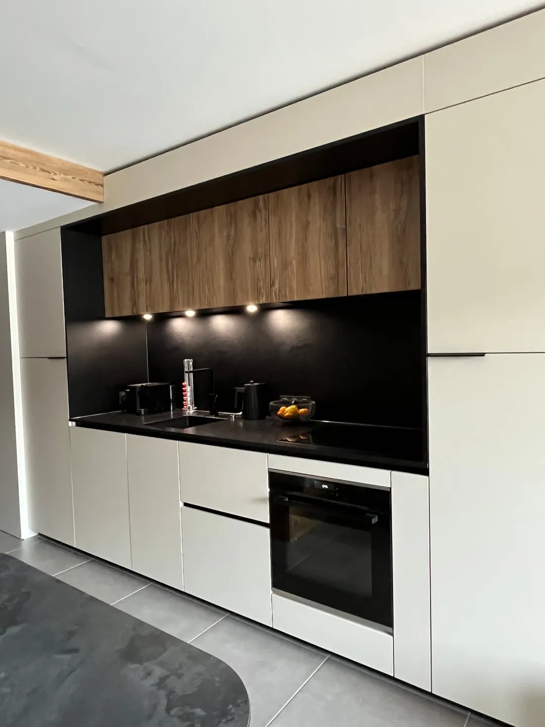 Cuisine linéaire toute hauteur à façades sable sans poignées, plan de travail et crédence noirs, meubles hauts en bois foncé éclairés par des spots encastrés