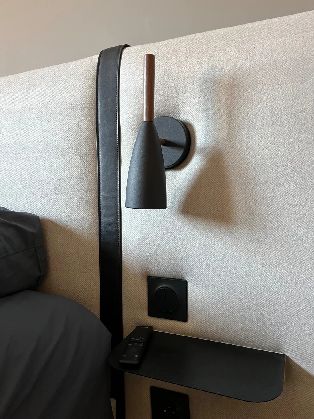 Détail de la tête de lit : applique de chevet en noyer et métal noir fixée sur le tissu chevronné écru, sangle de cuir noir tombant à côté, interrupteur noir et tablette de chevet en métal noir