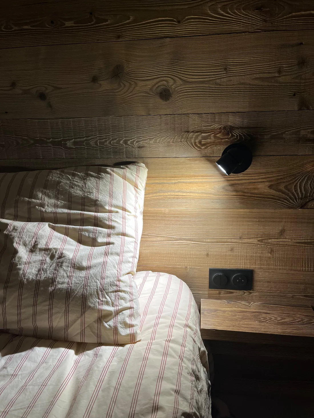 Gros plan sur une liseuse orientable noire fixée au lambris de bois ancien, au-dessus d'un oreiller en coton rayé rouge et blanc