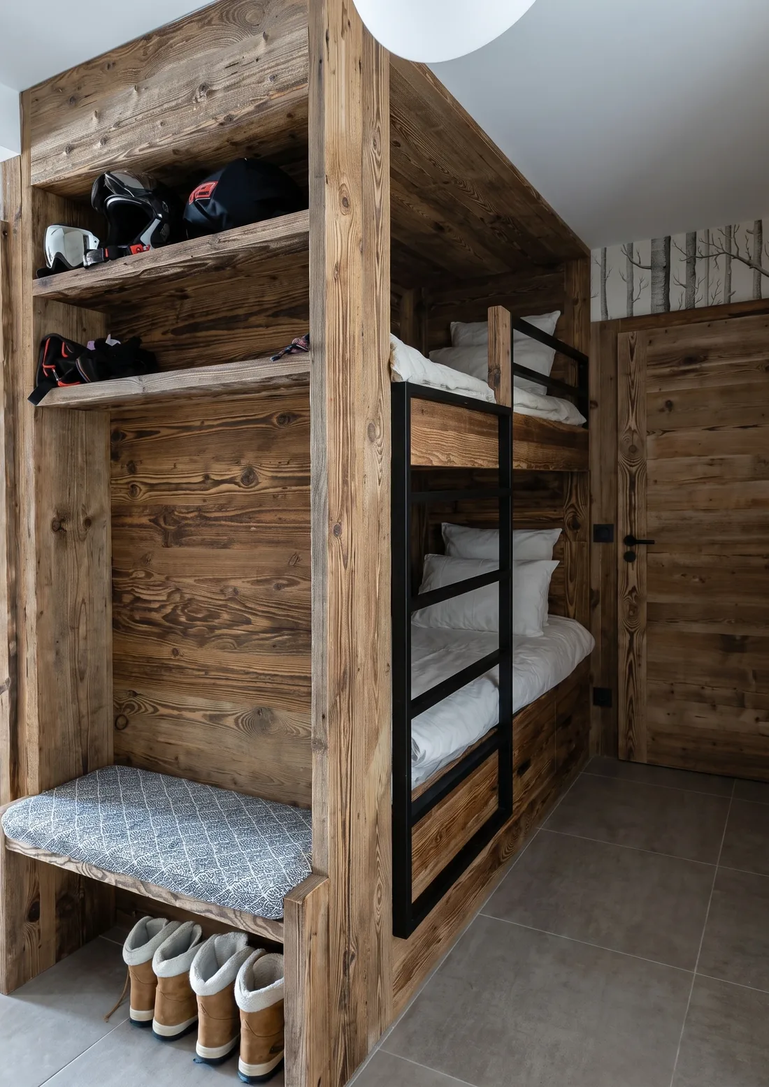 Bloc de lits superposés construit sur mesure en vieux bois : échelle en métal noir, liseuse encastrée dans le bardage, étagères hautes garnies de casques et de sacs de ski, banc d'assise en bas