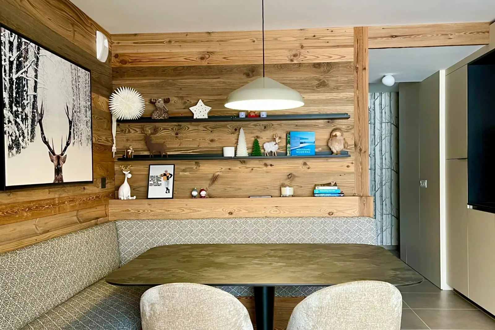 Coin repas de l'appartement de chalet : banquette d'angle sur mesure en vieux bois garnie d'un tissu à losanges gris, table ovale à plateau bronze, murs en bois de récupération et grande photographie d'un cerf sous la neige