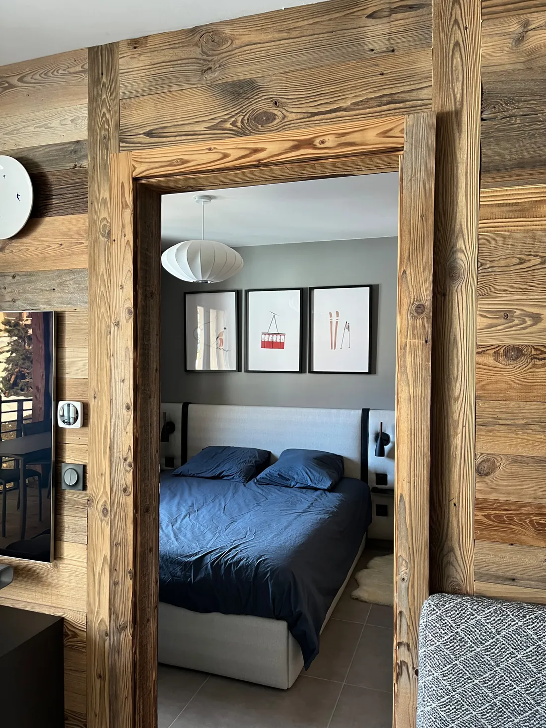 Ouverture encadrée de vieux bois entre le séjour et la chambre : on aperçoit le lit en linge bleu nuit, trois affiches de ski encadrées et une suspension boule blanche plissée