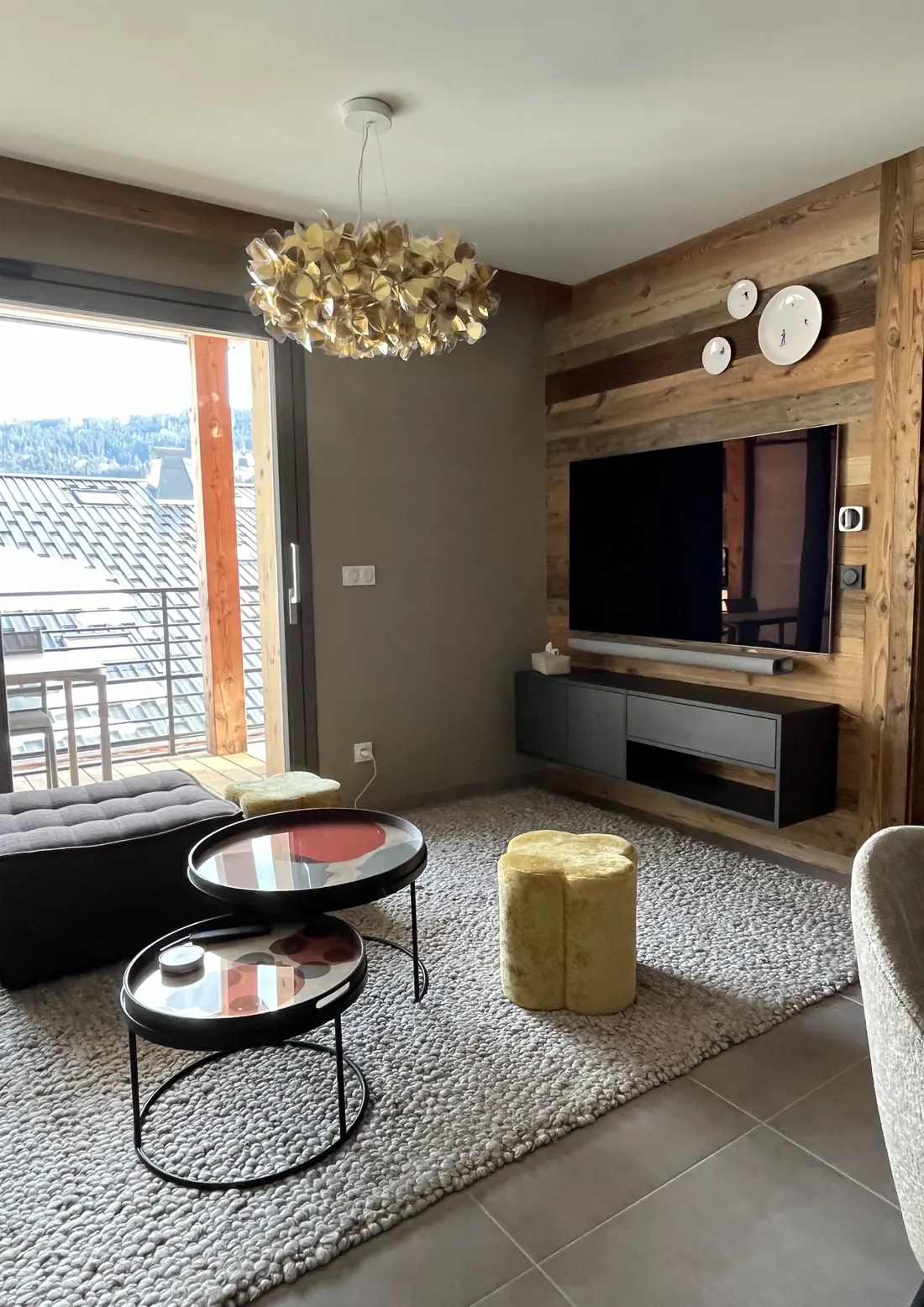 Séjour de l'appartement : mur habillé de vieux bois avec téléviseur encastré et meuble suspendu noir, tables gigognes rondes, pouf en velours jaune, tapis de laine bouclée et baie vitrée ouvrant sur les toits et la forêt
