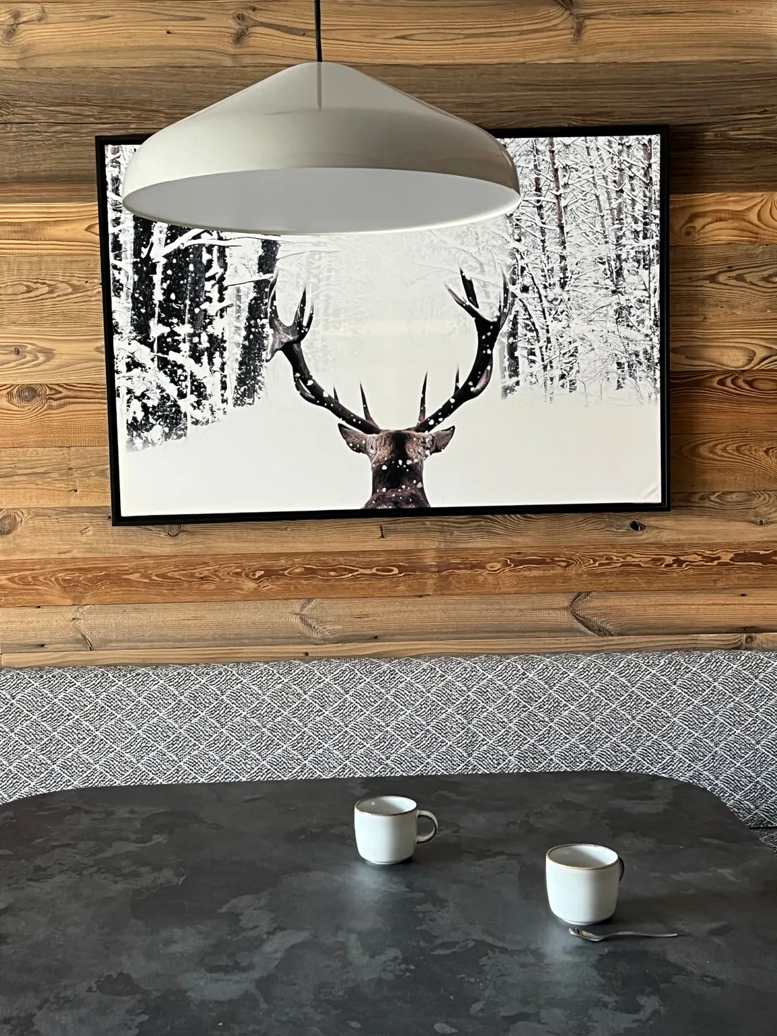 Détail du coin repas : grande photographie encadrée d'un cerf dans une forêt enneigée sur le mur de vieux bois, suspension blanche en dôme et plateau de table en pierre bronze