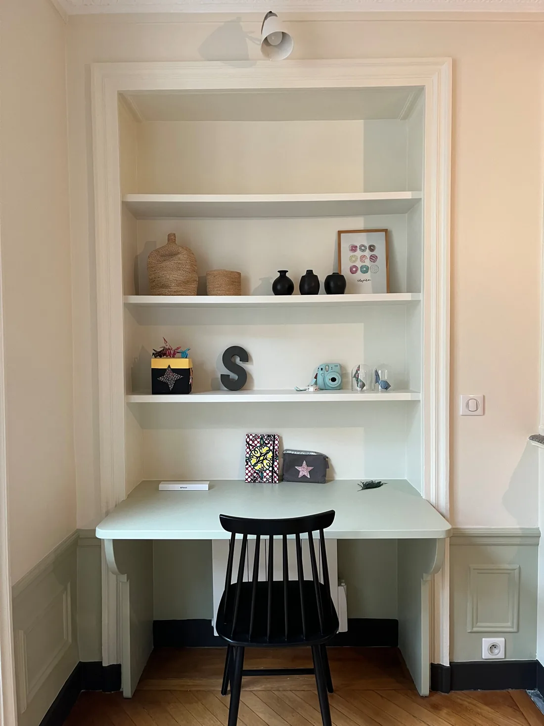 Le bureau en niche : plateau vert d'eau intégré dans l'épaisseur du mur, étagères garnies d'objets et d'une lettre S, chaise noire à barreaux et applique en hauteur