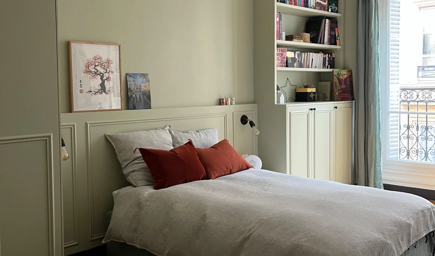 La chambre réalisée : lit aux coussins terracotta contre la tête de lit moulurée, placards vert tilleul à gauche, bibliothèque et fenêtre à droite