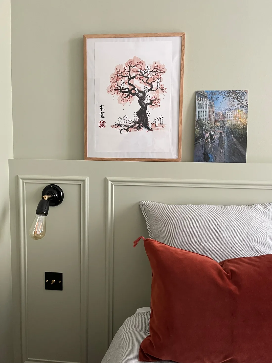 Détail de la tête de lit moulurée peinte en vert tilleul : applique noire à ampoule nue fixée sur la moulure, oreiller écru et coussin de velours terracotta, estampe de cerisier en fleur encadrée au-dessus