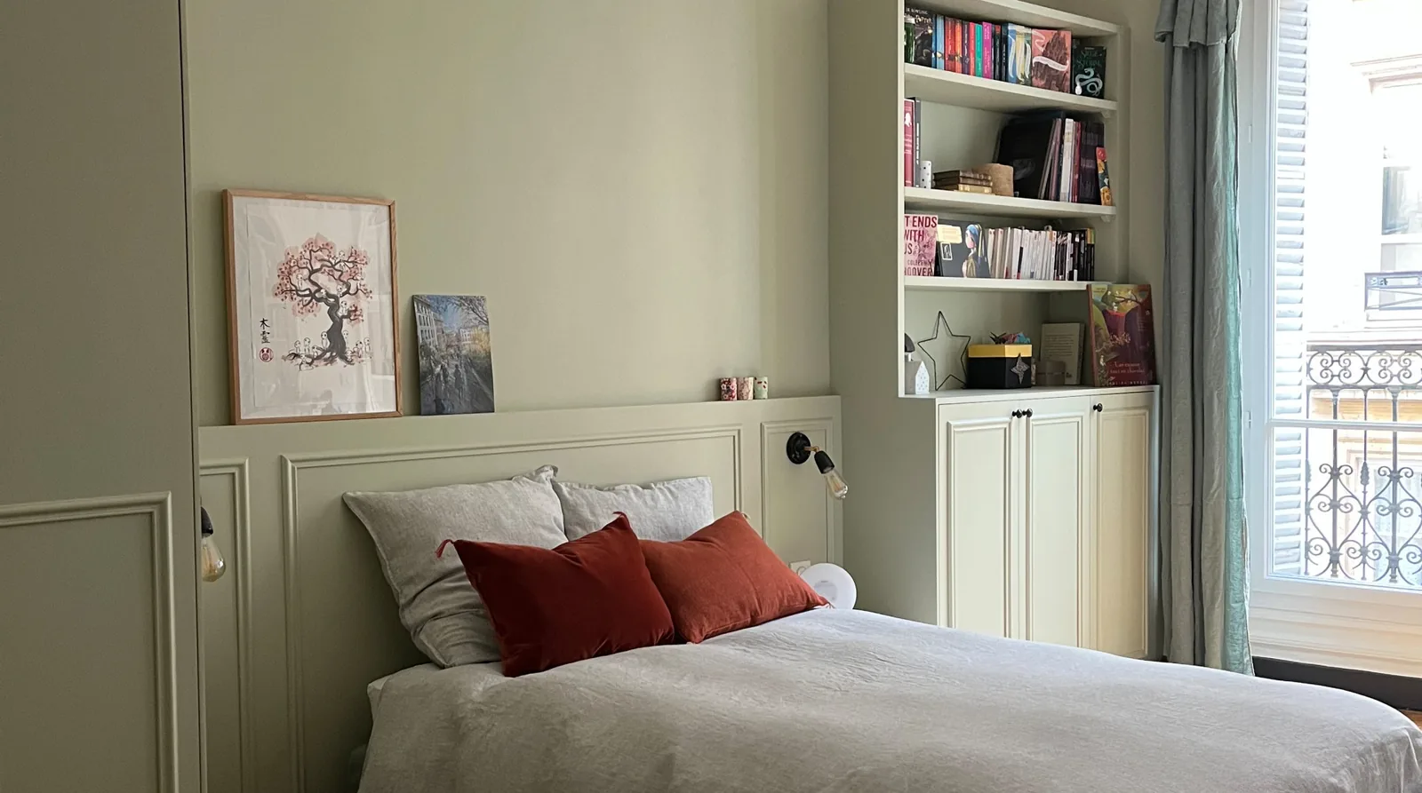 La chambre terminée : lit à coussin terracotta sous une suspension de rotin, bibliothèque sur mesure vert tilleul à portes moulurées près de la fenêtre, corniche ancienne et parquet point de Hongrie