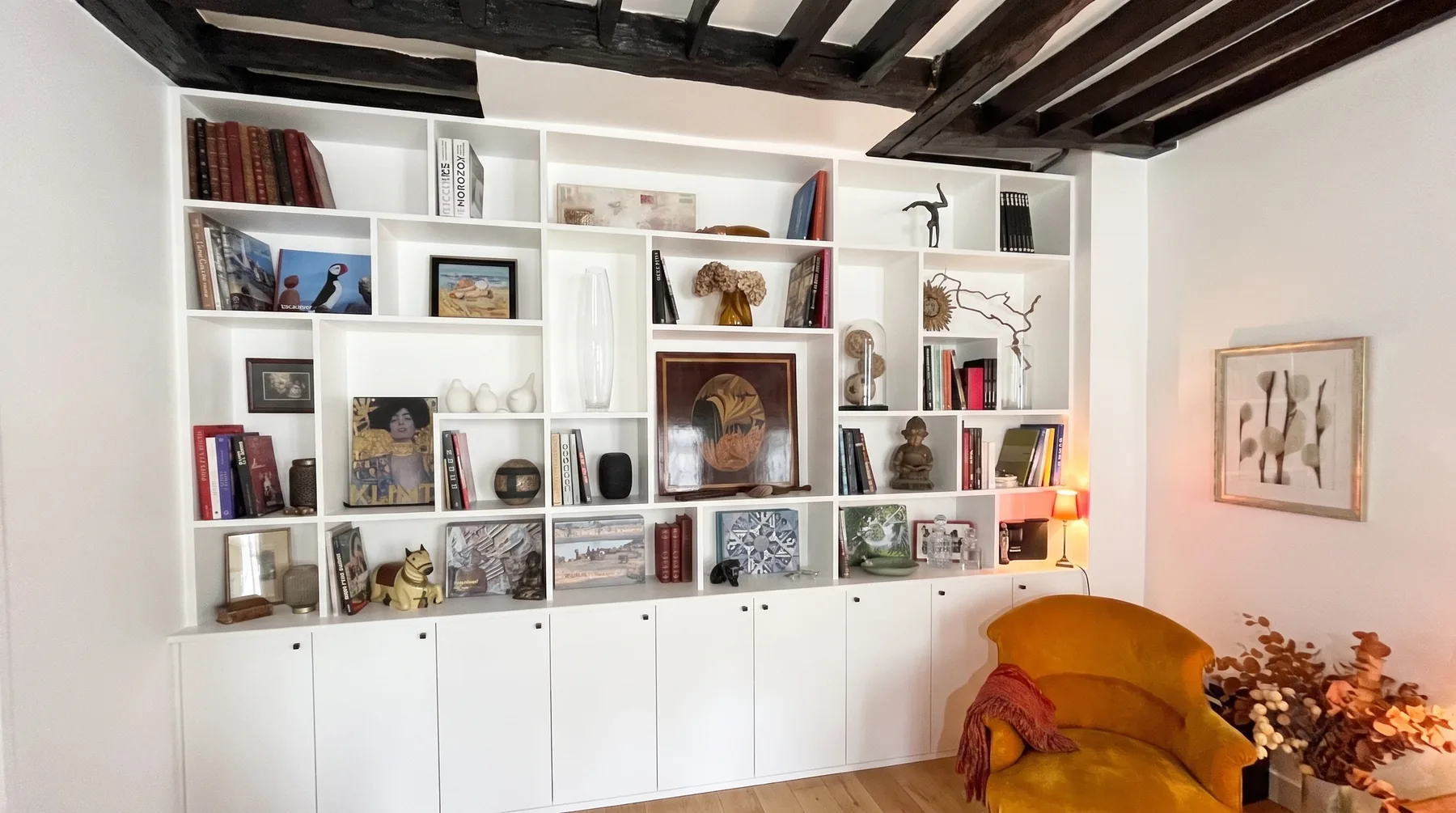 Bibliothèque sur mesure blanche occupant tout le mur du fond du salon : cases de tailles inégales garnies de livres, de cadres, de céramiques et de souvenirs de voyage, portes pleines en partie basse, sous un plafond à poutres apparentes sombres