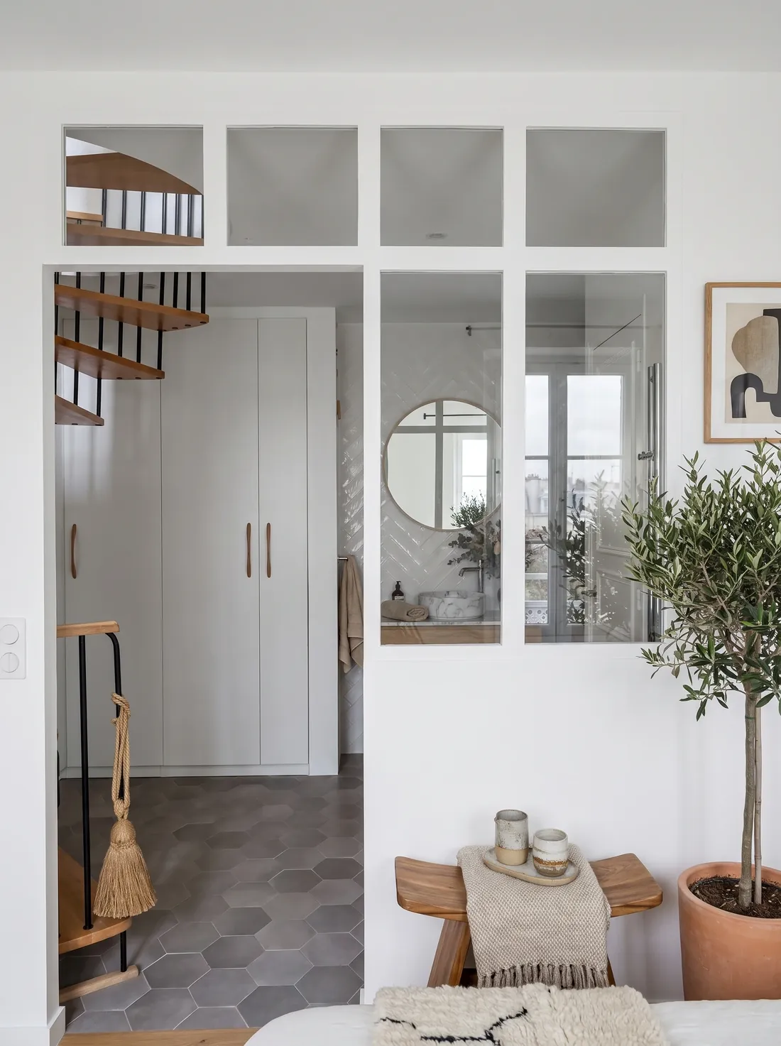 Cloison vitrée sur mesure séparant la chambre de la salle d'eau : impostes fixes en partie haute, dressing à portes lisses et poignées de cuir sur la gauche, escalier tournant en bois et métal noir, sol en carreaux hexagonaux gris, et au fond la vasque et le miroir rond de la salle d'eau