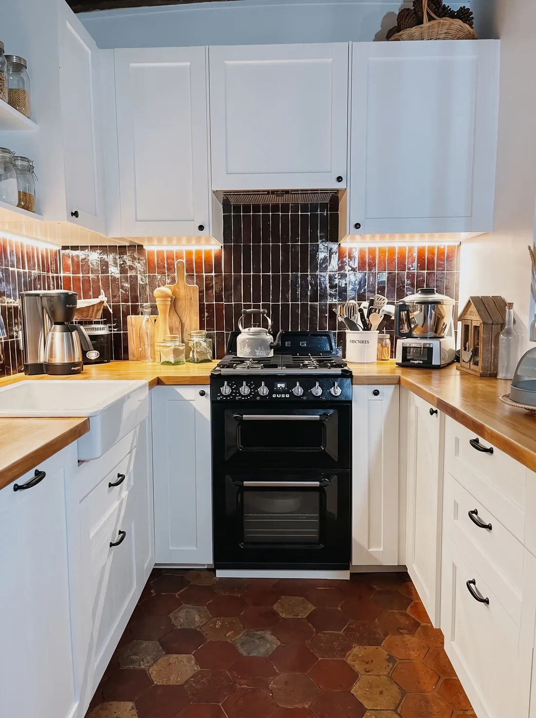 Cuisine du duplex : crédence de zelliges bruns brillants posés en verticale, plan de travail en bois massif, façades blanches à cadre et poignées noires, évier de céramique à tablier, piano de cuisson noir et sol en tommettes hexagonales anciennes