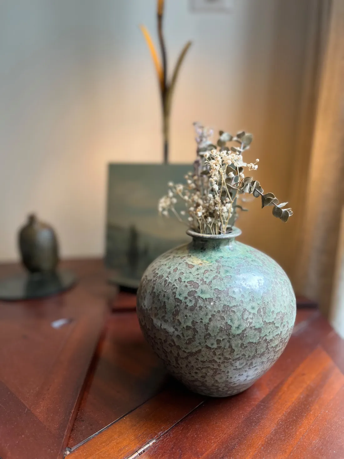 Détail : vase boule en grès émaillé vert mousse garni de fleurs séchées et d'eucalyptus, posé sur une commode d'acajou devant une petite marine encadrée