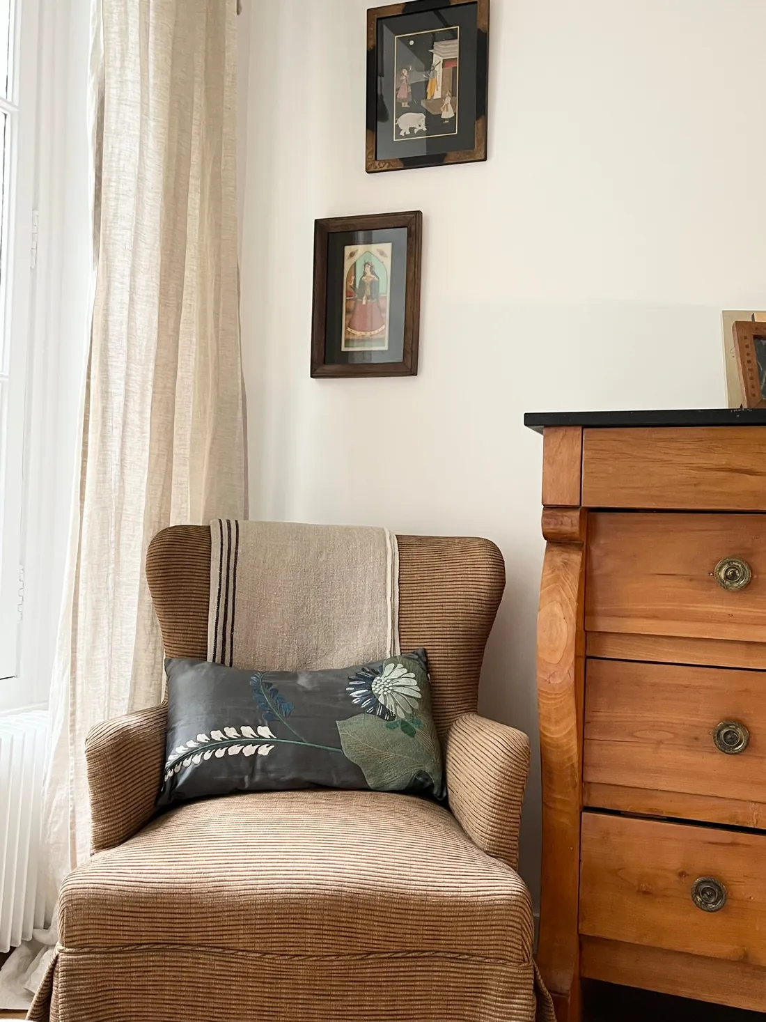 Fauteuil ancien retapissé d'un velours côtelé fauve, coussin brodé d'un chardon, jeté de lin rayé, à côté d'une commode de merisier à dessus de marbre noir et de deux miniatures indiennes encadrées