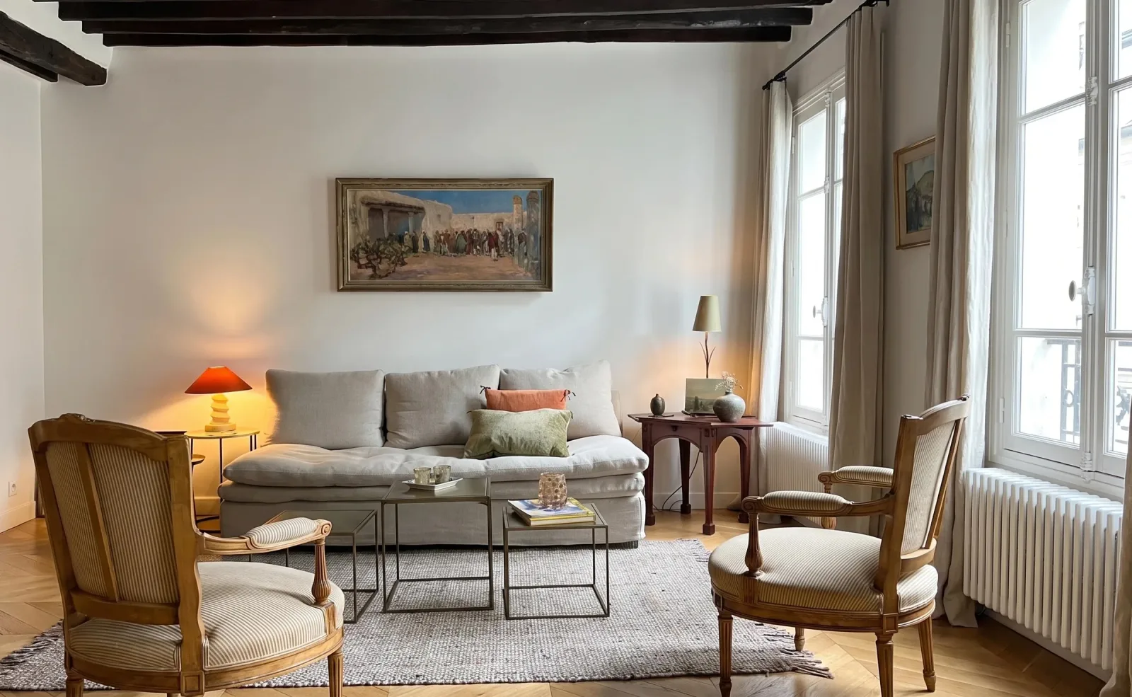 Salon du duplex : poutres anciennes peintes en brun sombre au plafond, canapé de lin écru, deux fauteuils cannés à rayures, parquet point de Hongrie et deux fenêtres à petits bois ouvrant sur la rue