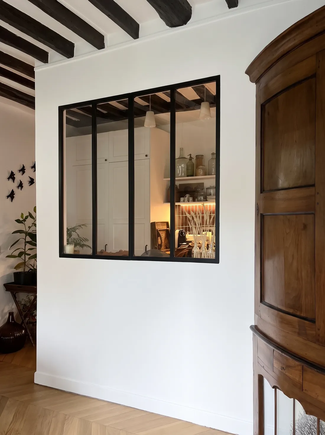 Verrière à châssis noir ouverte dans la cloison de la cuisine, vue depuis l'entrée : on aperçoit les étagères, les bocaux et les suspensions de la cuisine ; à droite une armoire ancienne en merisier, au sol un parquet point de Hongrie