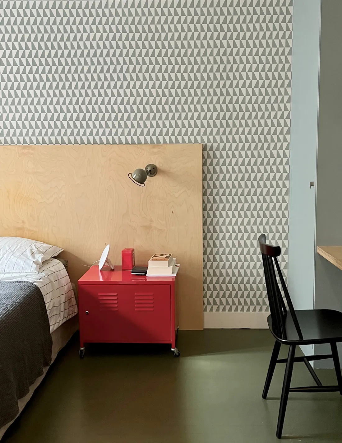 Chambre d'enfant : papier peint géométrique gris vert, tête de lit en contreplaqué de bouleau brut, applique d'atelier kaki et chevet en casier métallique rouge sur roulettes