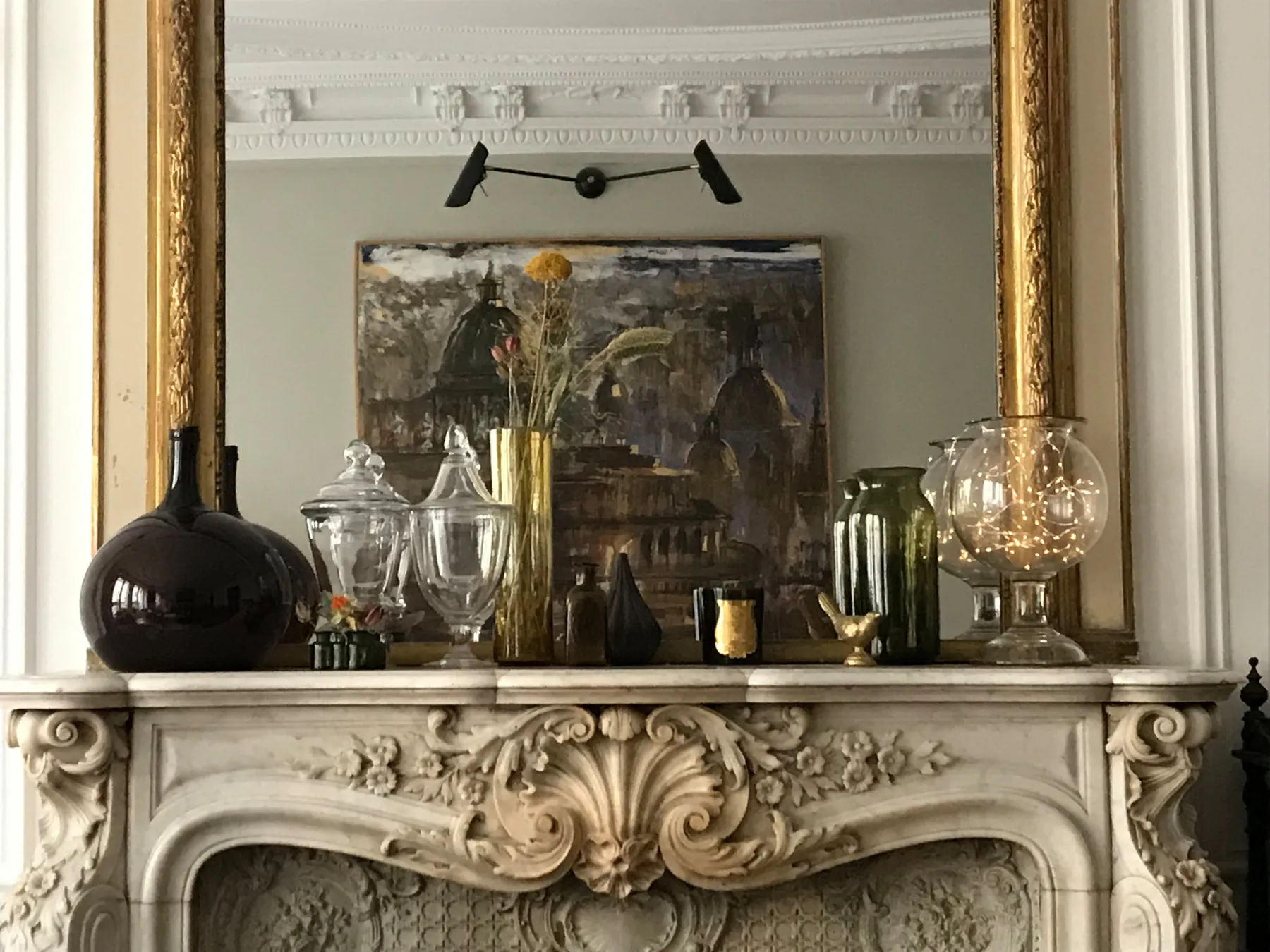 Cheminée de marbre blanc sculptée d'une coquille et de fleurs, surmontée d'un trumeau doré ; sur le manteau, une collection de verreries anciennes et de dames-jeannes