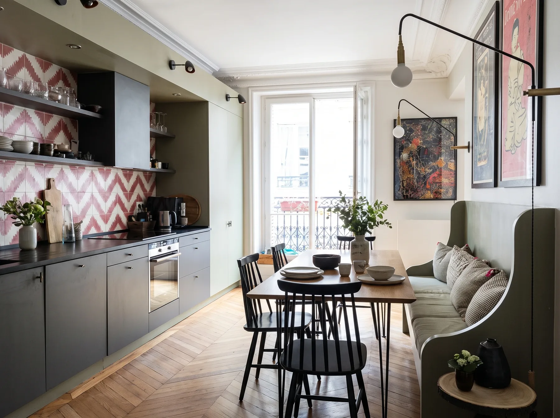 Vue d'ensemble de la cuisine et du coin repas : cuisine linéaire gris anthracite, table et chaises noires, banquette kaki, rosace au plafond et porte-fenêtre sur balcon