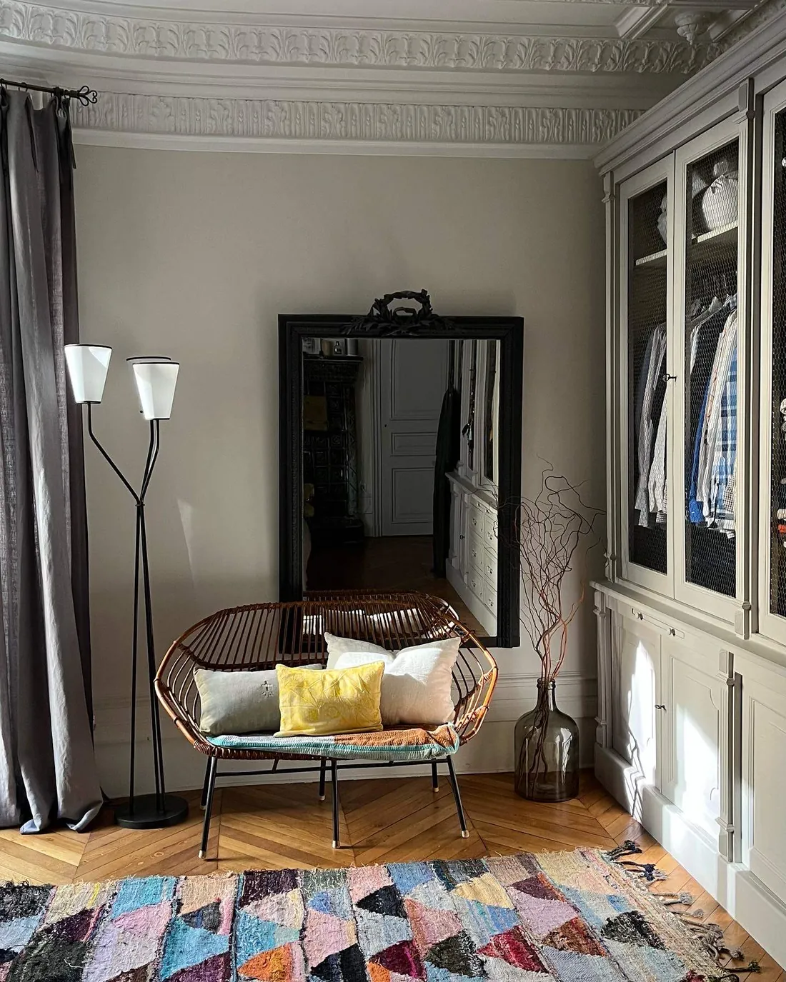 Armoire encastrée toute hauteur peinte en gris vert, portes garnies de grillage laiton laissant voir le linge, banquette en rotin vintage et grand miroir noir