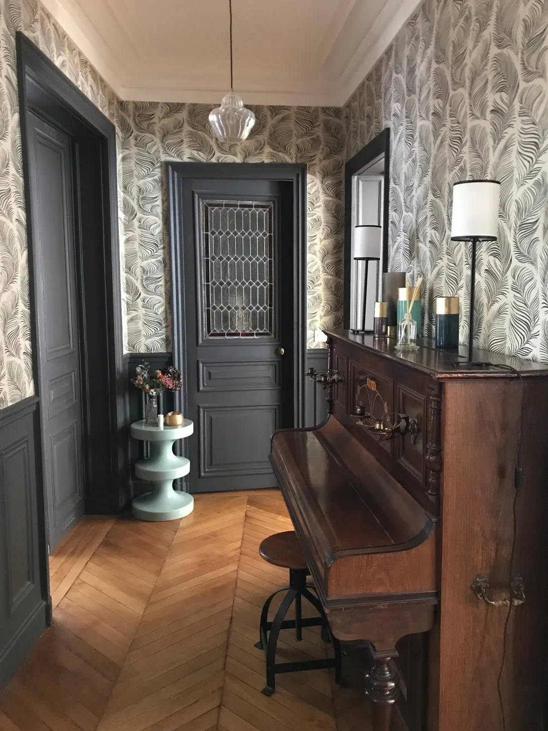 Entrée tendue d'un papier peint à grandes feuilles noires et blanches, portes et soubassements peints en gris ardoise, porte ancienne à vitrail plombé et piano droit en bois